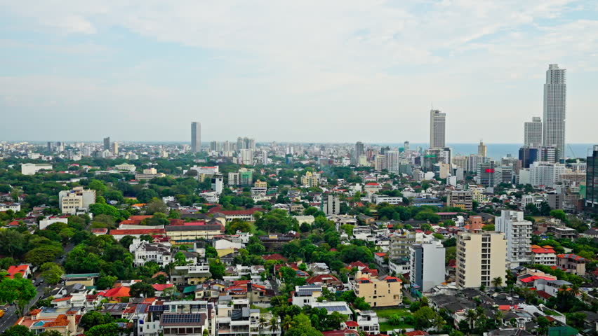 High angle view across Colombo, Sri Lanka, with clusters of modern high-rise towers dominating the skyline above dense low-rise neighborhoods, tropical greenery and the distant ocean horizon.