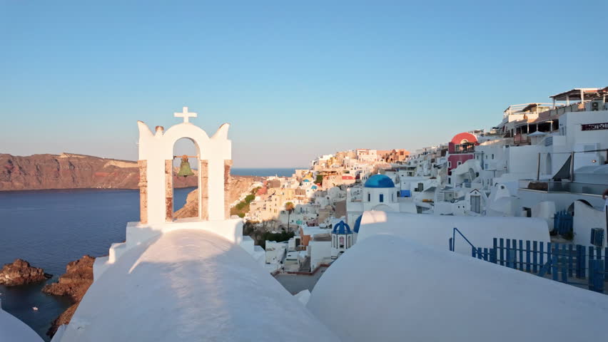 Church bell tower and white domes overlook a cliffside village and calm sea in warm sunset light, classic Santorini skyline view.
