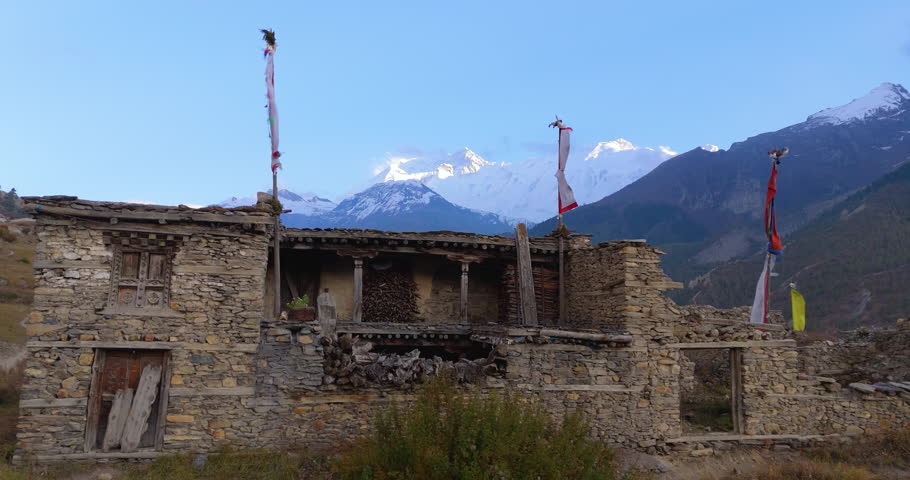 Annapurna Circuit, Manang, Nepal, drone shot of an old stone masonry home with wooden poles and prayer flags set against snow-covered mountain peaks under a clear blue sky.
