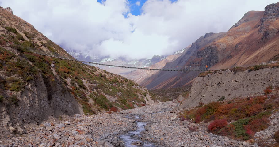 Annapurna Circuit, Manang, Nepal, push-in drone shot along a high suspension bridge over a mountain gorge toward trekkers with snow-capped Annapurna peaks in the background