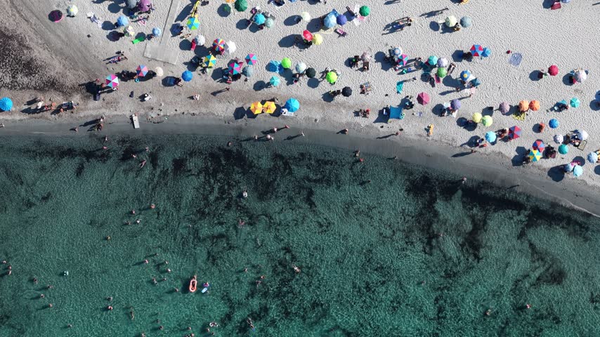 Aerial view of colorful beach umbrellas on the sand and people swimming in the clear turquoise sea in Sardinia, Italy.