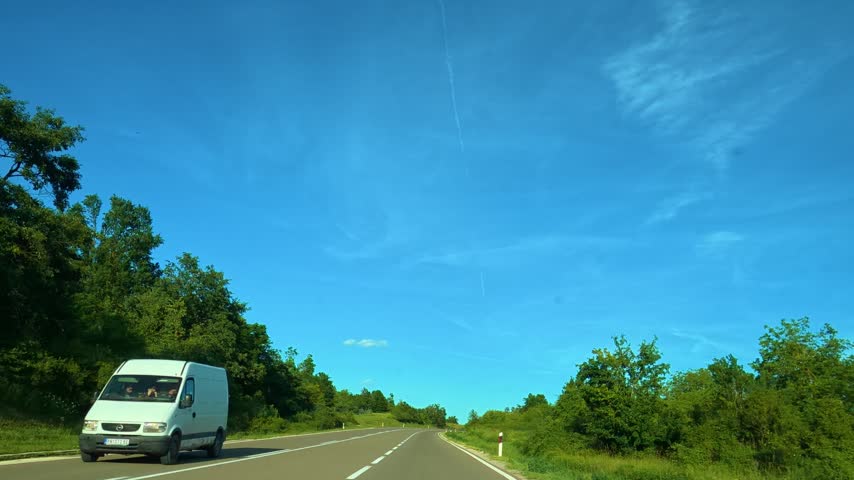 White van drives on open road under bright blue sky. Road curves gently through green trees and fields. Sky shows wispy clouds and distant mountains. Location: Mount Rtanj Area. Balkans, Serbia