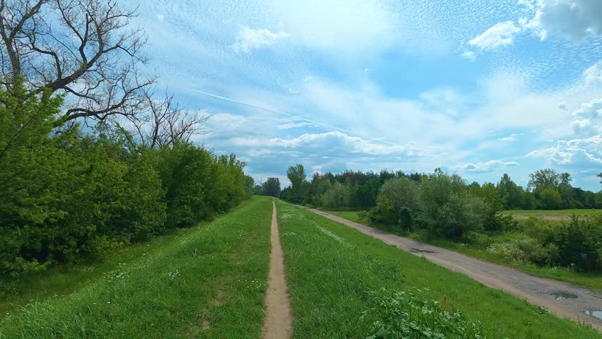 Green riverside path leading through rural embankment under sky. Bicycle POV.