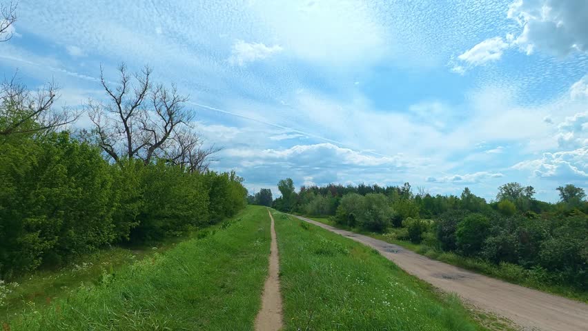 Rural trail stretching along green embankment beside dirt road. Bicycle POV.