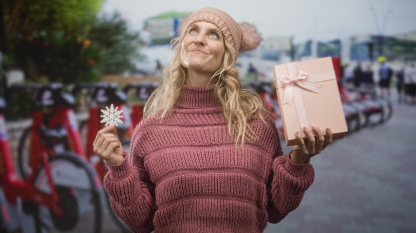 Woman holding a pink gift box and a snowflake ornament while looking up at a street bike station in winter; holiday joy.