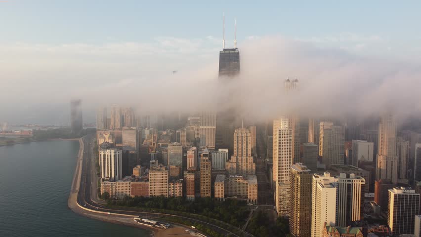 Aerial view of the Chicago skyline covered by low clouds and fog, with skyscrapers emerging above the mist and Lake Michigan along the waterfront.