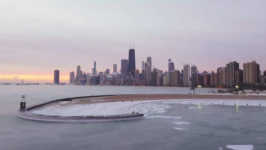 Wide view of the Chicago skyline and frozen Lake Michigan during a polar vortex, with ice along the shoreline and steam rising from the water at winter sunrise.