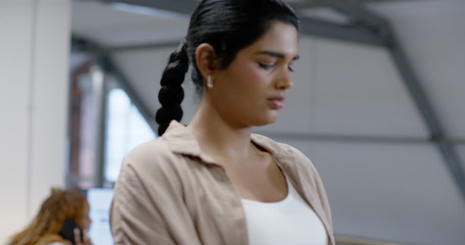 Indian woman standing in studio by railing, hearing comment and writing in notebook with metal pen. Portrait, focus, workspace, professional, modern, creative, monitors