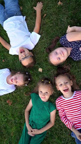 Top view of happy children lie in circle on grass in summer park look at camera and laugh. Team kids play outdoors. Childrens leisure. Vertical shot