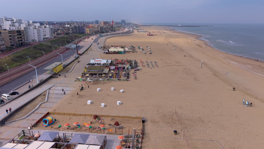 An aerial drone video shot providing a high-angle, wide perspective looking along the coastline and sandy beach at Scheveningen in The Hague, South Holland, Netherlands. The video frame features beachfront pavilions, outdoor restaurant terraces with sun loungers, giant white dice art sculptures on the sand, and a kids