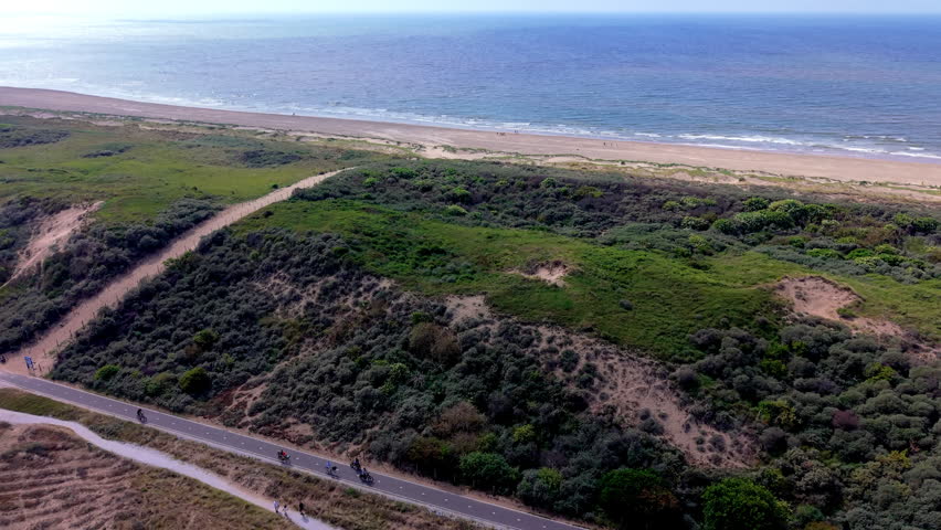 An aerial drone video shot providing a high-angle perspective over the coastal sand dune landscape and bicycle path near Katwijk aan Zee in South Holland, Netherlands. The video frame features several cyclists riding along a paved asphalt cycle path winding beneath lush green dunes and coastal vegetation. Above the dune hills, a sandy beach stretch and bright blue North Sea waters extend to a clear horizon under a sunny summer sky. High-definition video footage captures Dutch cycling culture, coastal dune conservation, seaside recreation, and popular European travel destinations on a bright daylight afternoon. The clip provides versatile stock video content for active lifestyle videos, cycling travel vlogs, nature documentaries, and European tourism promotions.