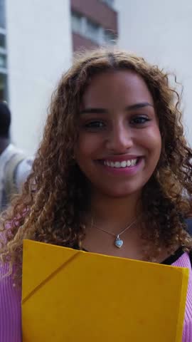 Vertical. Beautiful young african american woman with afro hair smiling and looking at the camera, standing in front of the university building with other students in the background on the stairs