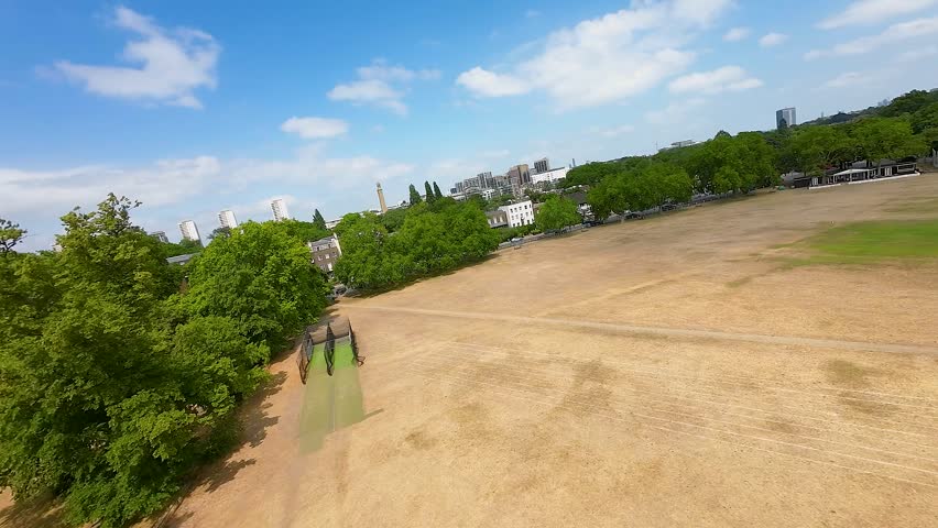 FPV aerial view of the expansive green space at Richmond Green with surrounding trees and historic buildings under a blue sky in Richmond, England, United Kingdom.