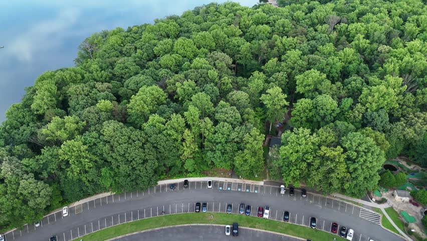 Cinematic aerial drone footage capturing a beautiful summer landscape in Burke, Virginia. The shot begins over a paved parking lot and slowly tilts up, gliding over a lush green forest canopy to reveal an expansive, tranquil lake. The glassy water beautifully reflects the sky. A fishing pier and distant shorelines become visible. This scenic establishing shot is perfect for projects highlighting parks, environmental conservation, and outdoor recreation.