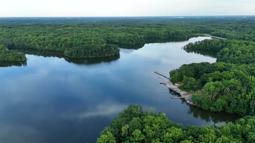 Cinematic aerial drone footage sweeping over a serene lake surrounded by dense, lush green summer forests in Burke, Virginia. The expansive high-angle view reveals a peaceful recreational park environment, featuring a wooden dock, concrete boat launch, and small kayaks paddling on the calm water that reflects the sky above. Ideal for nature, outdoor recreation, environmental, and regional community lifestyle content.