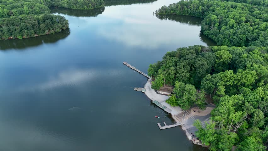 Cinematic aerial drone footage moving slowly over a serene lake, reflecting the blue sky and clouds above. The camera pushes forward toward a recreational boat launch area featuring wooden docks, a concrete ramp, and a shoreline facility. Dense, lush green summer trees surround the water