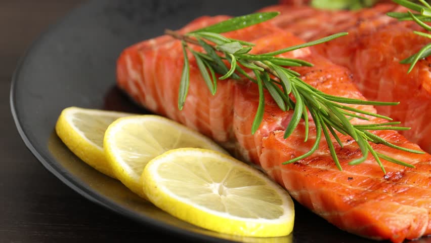 Pieces of grilled salmon with thyme, lemon and salad on wooden table, closeup. Camera moving right