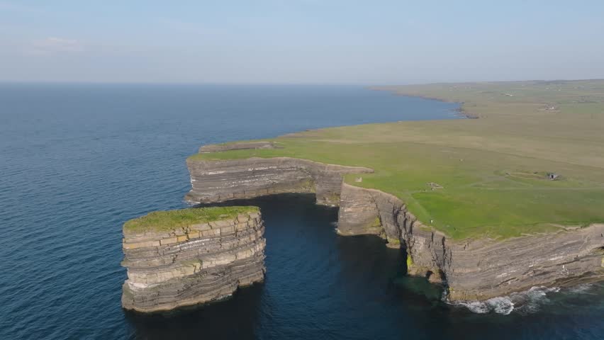 Layered rock column Dun Briste stands off a flat green headland above calm Atlantic water, viewed from a high circling drone.