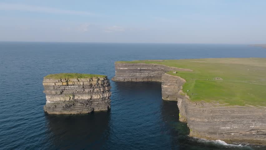 Layered rock column Dun Briste stands off a flat grassy cliff top above calm Atlantic waters in bright daytime light.