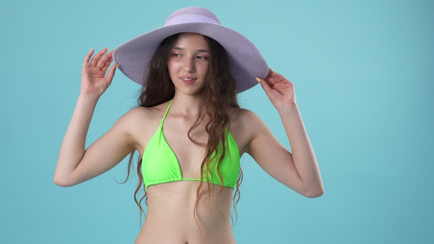 A smiling young woman wearing a large purple sun hat, white sunglasses, and a green bikini, posing playfully against a blue background.