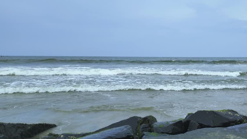 Seawater rolling onto wet sand with foam hitting textured stone barriers under overcast sky at peaceful seaside coastline nature scene.