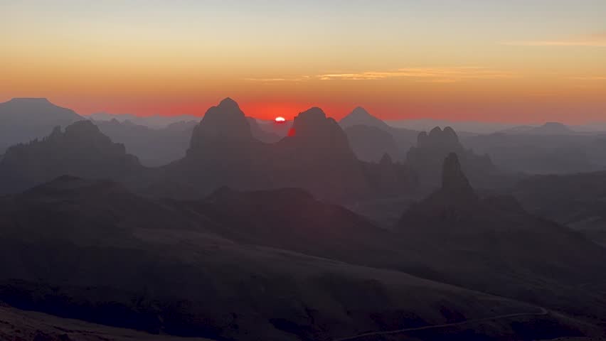 A stunning panoramic view of the sun rising behind the dramatic, jagged rock formations of the Hoggar Mountains in the Sahara Desert, Algeria.