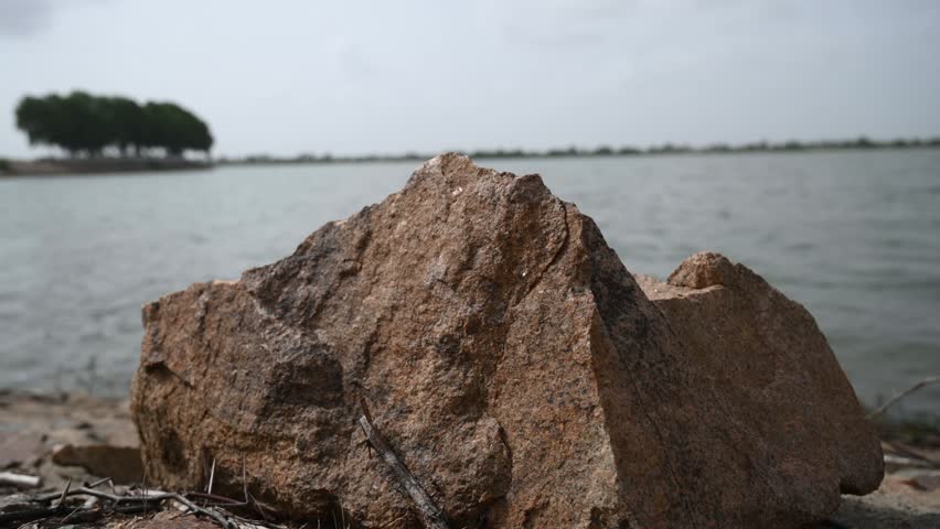 Large Natural Rock on Lakeshore with Calm Water and Distant Green Landscape, Scenic Rural Nature and Peaceful Outdoor Environment 4K Footage.tharparkar
