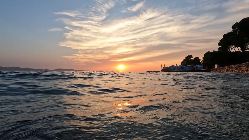 Scenic sunset over Adriatic sea with warm orange sky, rippling water, and coastal shoreline. Peaceful Croatian seaside scene, tourism, and Mediterranean lifestyle.