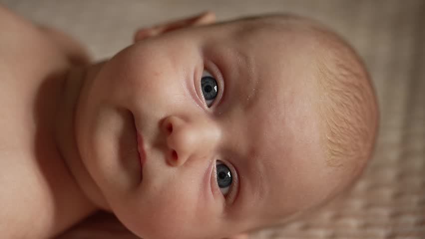 Close up portrait of a cute newborn baby with big blue eyes looking directly into the camera. An adorable infant with soft skin making different facial expressions while lying down