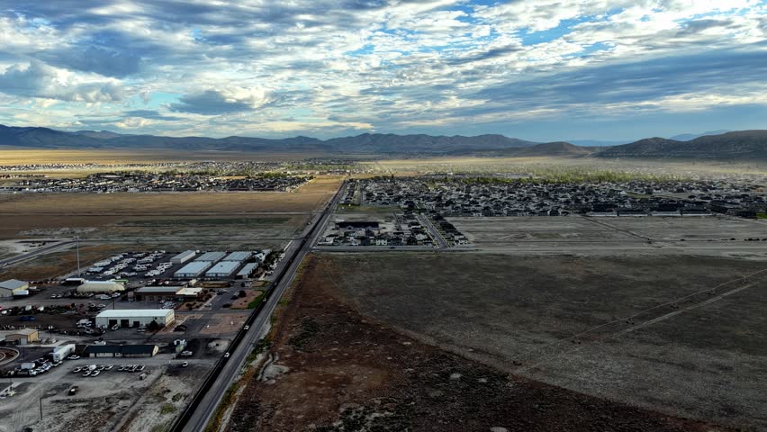 Stunning drone video tilting down over Eagle Mountain, Utah, shows rapid suburban growth, new residential construction, sunlit clouds, wide roads, and scenic mountain backdrop.
