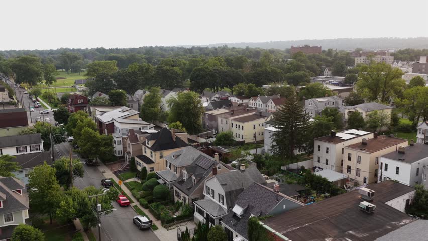 Dense row of gabled colonial homes lines tree shaded street in Orange, New Jersey, parked cars and distant green playing fields stretch beyond hazy town rooftops nearby. Aerial drone rising shot