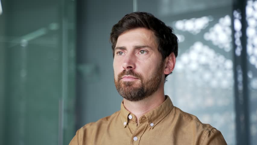 Close-up portrait of a smiling bearded mature businessman in a shirt looking at the camera in a modern glass office. Confident professional posing with a friendly expression in a corporate workplace.