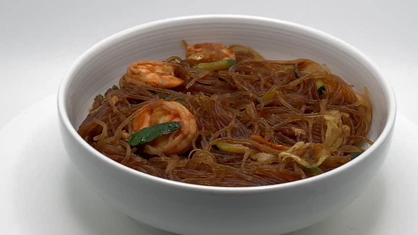 Traditional stir fried glass noodles with shrimp and vegetables served in a white bowl against a plain white background.
