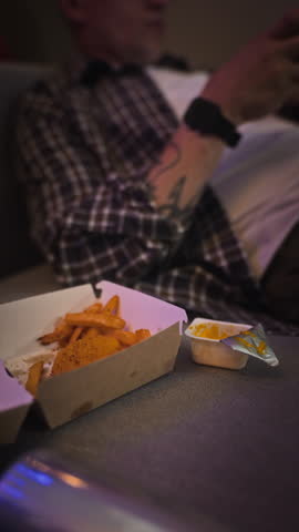 Vertical video of Caucasian young adult man dipping fried chicken in sauce and eating while staring at screen. Clip represents screen addiction, unhealthy lifestyle, and distracted eating habits