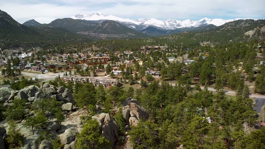 Drone footage flying over pine forest with large rocks, showing town buildings and snowy mountains in distance. Peaceful mountain village scenic view.