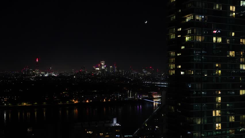 High-quality horizontal 4K night timelapse video capturing the crescent moon moving across the dark sky over the London skyline, framed by a lit residential skyscraper in the foreground