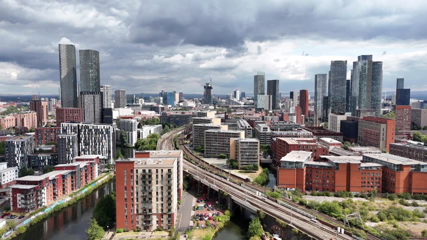 Tall skyscrapers and apartment buildings rise above city railway tracks in Manchester under a cloudy sky during the afternoon.