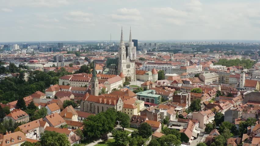Aerial cinematic footage captures sprawling cityscape of Zagreb with iconic cathedral towers rising above historic red rooftops on sunny day. Dynamic urban panorama showcases old town architecture.