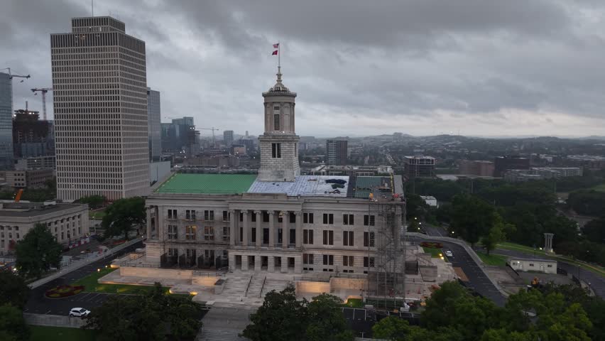 Side angle aerial drone footage of the Tennessee State Capitol tower in Nashville with an adjacent office building and winding roads around the grounds