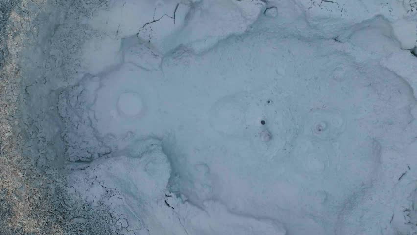 Cinematic overhead drone footage of geothermal activity. Bubbling grey mud pots and smoking volcanic vents. Textural ground patterns in Hverir Iceland. Dynamic natural thermal energy.