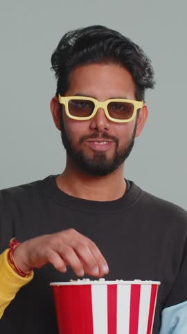 Excited Indian man in 3D glasses eating popcorn and watching interesting tv serial, sport game, film, online social media movie content. Hindu boy enjoying domestic entertainment on gray background
