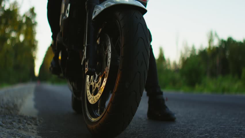 Sport motorcycle rider pauses on a quiet countryside road at sunset, highlighting front wheel, braking system, and powerful stance against blurred forest background