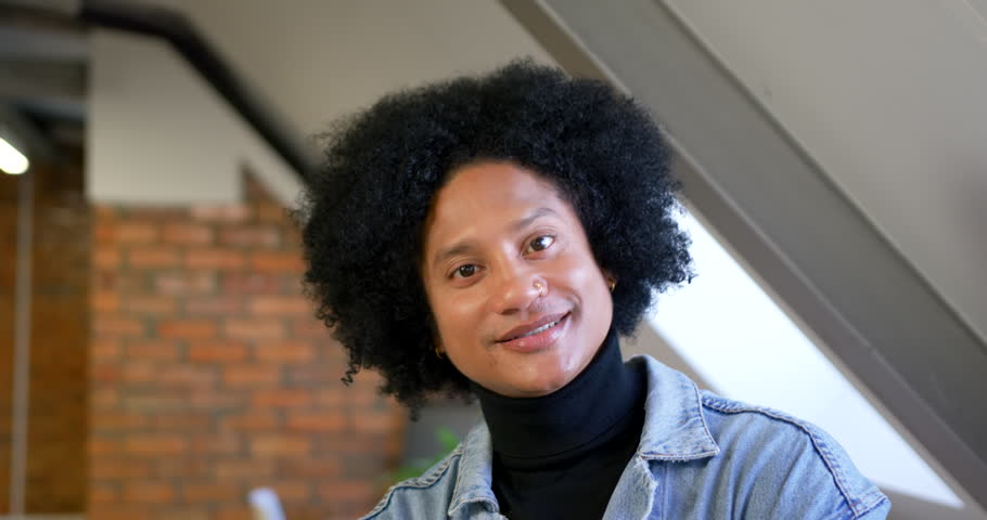 African American man in denim jacket noticing off-frame person, turning head right, returning smile. Loft, brick, piping, industrial, coworking, modern, gaze