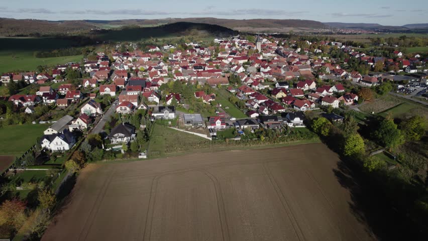 High angle aerial view of compact European town with church steeple, red roof houses, autumn trees, winding streets, and surrounding countryside under clear sunlight