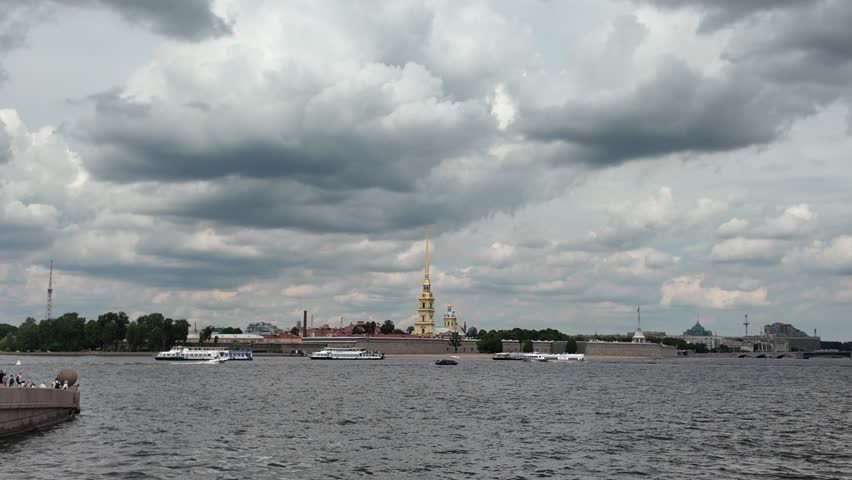 Panoramic view of the Peter and Paul Fortress and its iconic golden cathedral spire across the Neva River in Saint Petersburg, Russia. Passenger boats cruise along the river beneath spectacular dark summer clouds, creating a dramatic cityscape of one of the most famous landmarks in the historic center of Saint Petersburg.