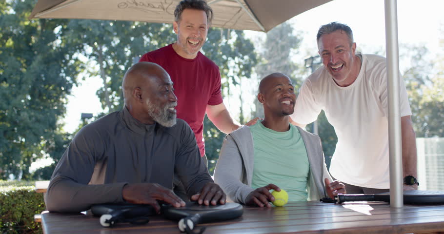 Diverse male friends gathering at picnic table, grabbing paddles and yellow ball for friendly game. Group, camaraderie, outdoors, park, shade, sunshine, casual