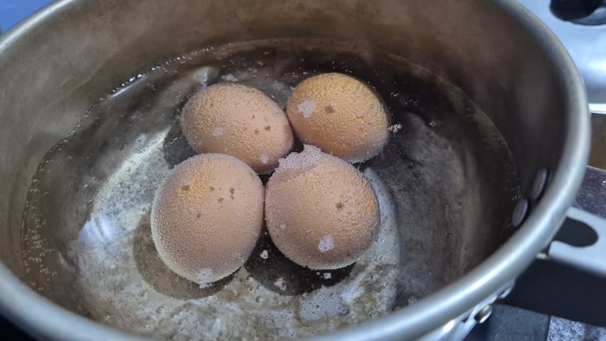 Close up top down shot of four brown eggs boiling in hot water with micro bubbles on shells inside a pot on a stove.
