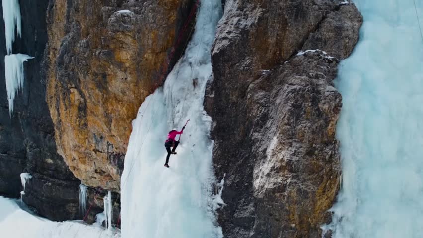 Aerial drone shot of an ice climber using axes and crampons to scale a steep, frozen ice fall attached to a rugged alpine rock cliff face.