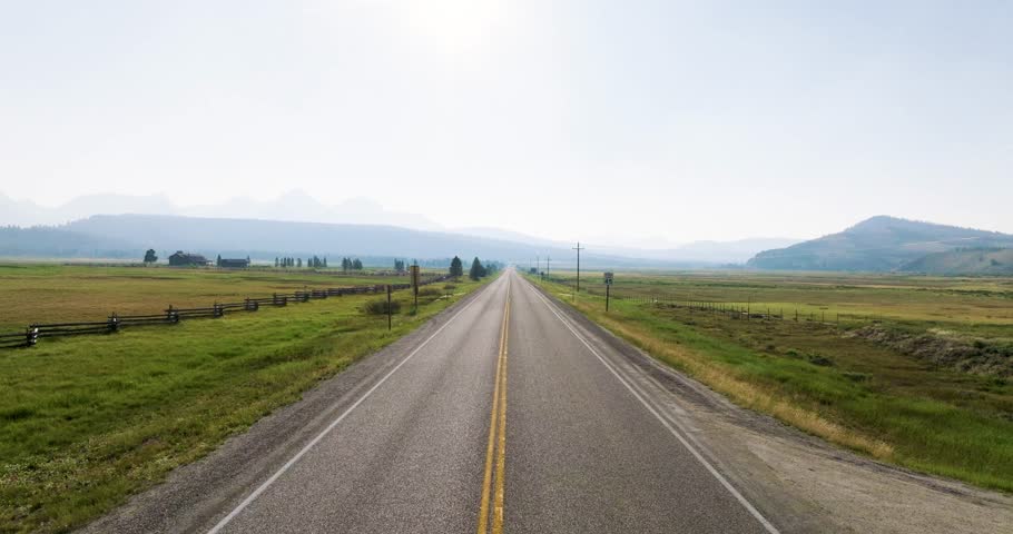 Rising shot of an asphalt road in stanley idaho with wide open fields on both sides. Distant mountains rise under a clear sky. Wide aerial view captures the endless countryside