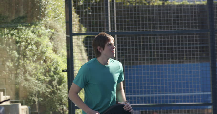 Adult male shifting, swinging black paddle, returning yellow ball approaching right at paddle court. Racquet, player, outdoor, glass, panels, tennis, movement